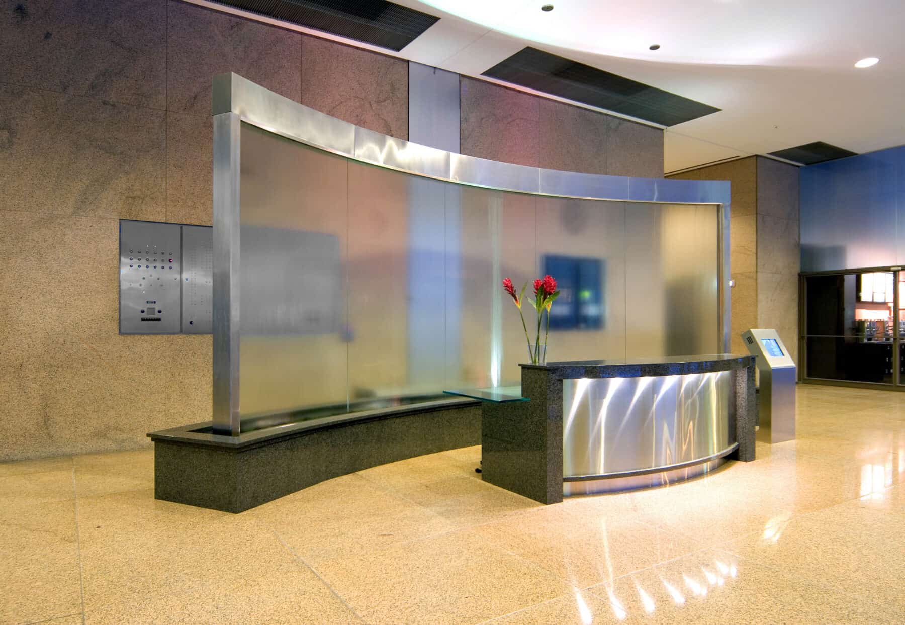 Custom Curved Glass Water Fountain Feature Behind Stone and Metal Reception Desk by Commercial Builder & General Contractor Structural Enterprises